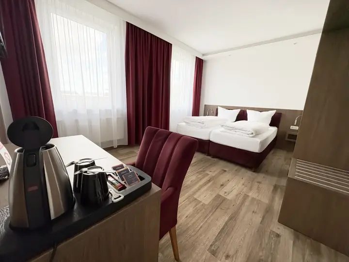 Doppelzimmer in Weinrot: Schreibtisch mit Wasserkocher und weinrotem Stuhl, dahinter das Bett